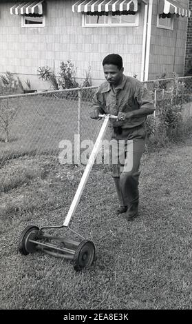 1960s, historisch, Mann mit einer kleinen Hand oder Push-Mäher mit einem Holzgriff, um das Gras auf seinem kleinen Stück Vorderrasen neben einem niedrigen Drahtzaun in einem Vorort der Stadt Chicago, USA schneiden. Stockfoto