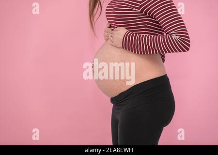 Der freiliegende Bauch einer jungen schwangeren Frau auf einem rosa Hintergrund. Seitenansicht und Nahaufnahme. Schwangerschaftskonzept. Stockfoto