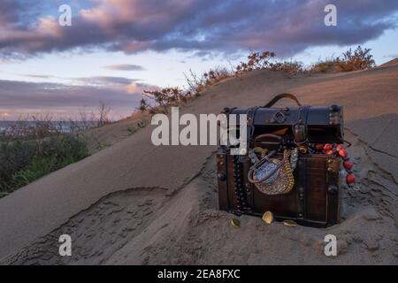 Schatzkiste am Strand bei Sonnenaufgang, Juwelen und Goldmünzen Stockfoto