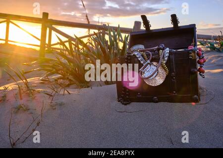 Schatzkiste am Strand bei Sonnenaufgang, Juwelen und Goldmünzen Stockfoto