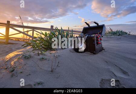 Schatzkiste am Strand bei Sonnenaufgang, Juwelen und Goldmünzen Stockfoto