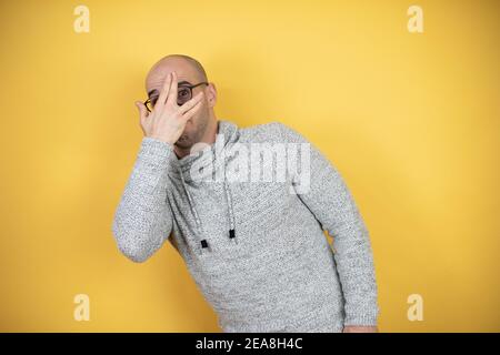 Junger kahler Mann mit Brille auf gelbem Hintergrund, der mit der Hand ins Gesicht und in die Augen schauend, mit peinlicher Expr durch die Finger schauend Stockfoto