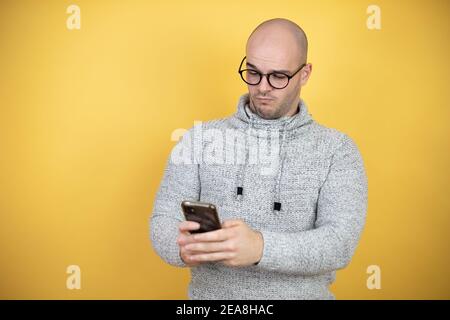 Junge Glatze Mann trägt Brille über gelben Hintergrund SMS mit Sein Telefon Stockfoto