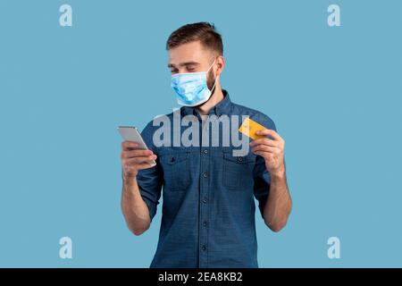 Kontaktloses Einkaufen während der Covid-Isolation. Porträt von Millennial Kerl in Gesichtsmaske hält Smartphone und Kreditkarte Stockfoto
