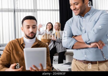 Lächelnde Geschäftsfrauen nehmen Selfie mit Smartphone in der Nähe von multiethnischen Kollegen auf Verschwommener Vordergrund Stockfoto