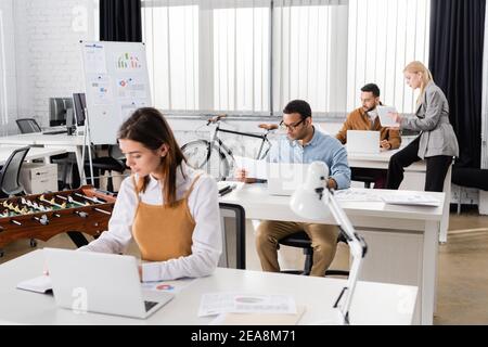 Indischer Geschäftsmann, der mit Papieren und Laptop in der Nähe multiethnischer Kollegen arbeitet Auf unscharfem Hintergrund Stockfoto