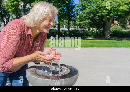 Reife Frau erfrischend aus Trinkwasser Brunnen am heißen Sommertag. Blonde Frau spritzt Wasser von Brunnen. Frisch durch die Hitze Stockfoto