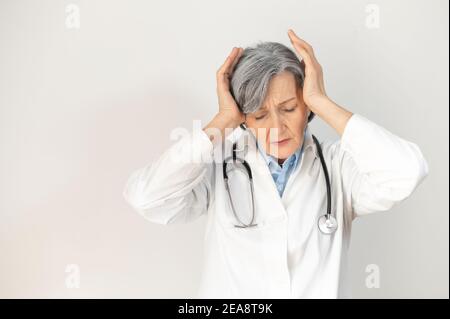 Gestresster älterer reifer Arzt mit einem Stethoskop in einem medizinischen Gewand, der Kopf in den Händen hält, überwältigt fühlt, erhielt schlechte verheerende Nachrichten Stockfoto