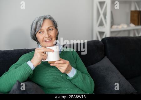 Ältere ältere grauhaarige Geschäftsfrau bekam ein kostenloses Getränk Angebot Kaffee Tee während in der Warteschlange warten, genießen heiße Getränke, sitzen im Flur Stockfoto