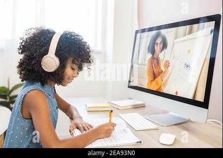 Ein cleveres multiethnisches Mädchen sitzt mit einem Kopfhörer am Tisch, studiert auf der Distanz, macht Hausaufgaben, schreibt an Arbeitsmappen, schaut sich Online-Kurse an Stockfoto