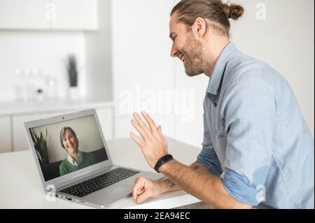 Ein Kerl winkt Hand, Begrüßung mit einer älteren Frau auf dem Laptop-Bildschirm, Student im virtuellen Gespräch mit älteren weiblichen Tutor, ein erwachsener Sohn ruft Stockfoto