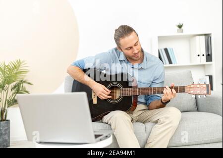 Ein junger Mann lernt Gitarre spielen selbst, beobachten Webinar, Online-Masterclass, Video-Tutorial. Ein Kerl hält akustische Gitarre und wiederholen Übungen Stockfoto