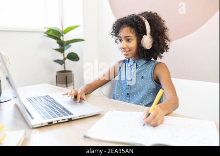 Ein hübsches lockiges afrikanisches Schulmädchen trägt einen Kopfhörer mit einem Laptop für das Lernen auf der Ferne, ein hübsches biracial Mädchen füllen Übungen Buch mit Stockfoto