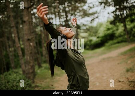 Hübsche junge Frau mit Kopfhörer, die ihre Arme im Wald vorpreading, weil sie draußen trainiert Stockfoto