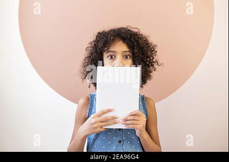 Ein ängstliches und schüchternes afrikanisches Schulmädchen bedeckt die untere Hälfte des Gesichts mit einem Buch und fürchtet, die Prüfung nicht bestehen zu können Stockfoto