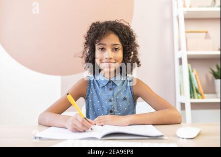 Webcam-Ansicht eines ziemlich lockig afrikanischen Schulmädchen trägt einen Kopfhörer, ein hübsches biracial Mädchen füllen Übungen Buch mit einem Stift beobachten Online-Video Stockfoto