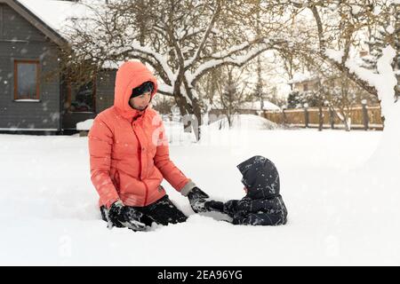 Junge Mutter und Sohn sitzen im tiefen Schnee und genießen Zeit zusammen an einem sonnigen Wintertag. Glückliches Familienkonzept. Stockfoto