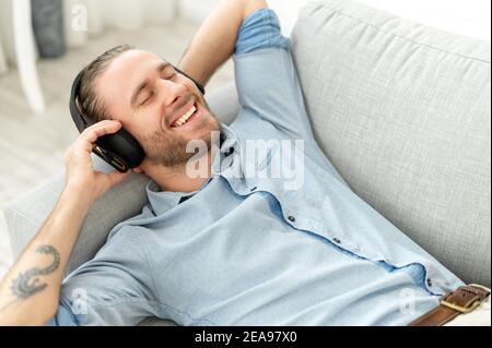 Zufriedener Kerl genießt Musik auf der Couch liegen. Ein lächelnder Hipster mit Kopfhörern, der Lieblingstitel hört, Hörbücher auf dem Sofa ausruhen Stockfoto