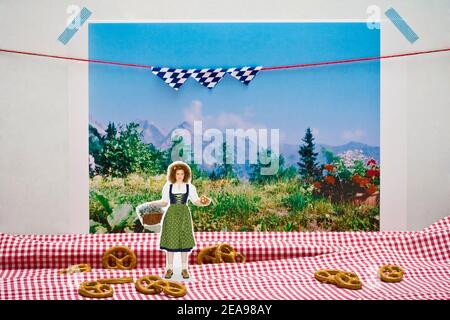 Stillleben Foto, Frau im Dirndl, mit Brezel in der Hand, Blumenkorb, rot-weiß karierte Tischdecke mit Brezeln als Hintergrund, Foto der bayerischen Landschaft als Hintergrund, bayerische Fahnen Stockfoto