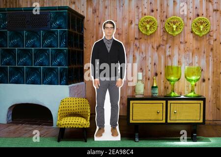 Fotocollage, Mann neben Sessel, vor Regal und Kachelofen, grüner Teppich, Gläser, gelb, Hirschgeweih an der Wand Stockfoto