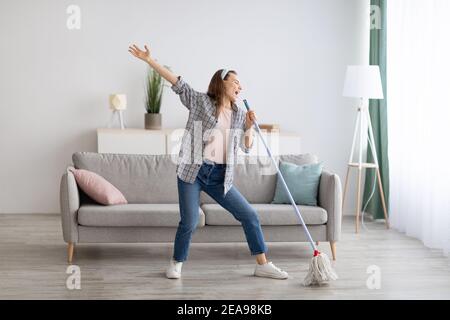 Glückliche Frau singen beim Waschen Boden im Wohnzimmer, mit Mopp als Mikrofon, Spaß während Hausarbeiten Stockfoto