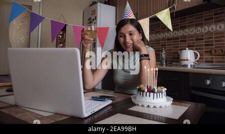 Frau feiert ihren Geburtstag durch Videoanruf virtuelle Party mit Freunden. Lits und bläst Kerze aus. Authentisch dekorierter Arbeitsplatz. Coronavir Stockfoto