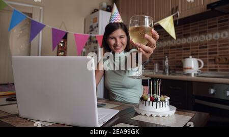 Frau feiert ihren Geburtstag durch Videoanruf virtuelle Party mit Freunden. Lits und bläst Kerze aus. Authentisch dekorierter Arbeitsplatz. Coronavir Stockfoto