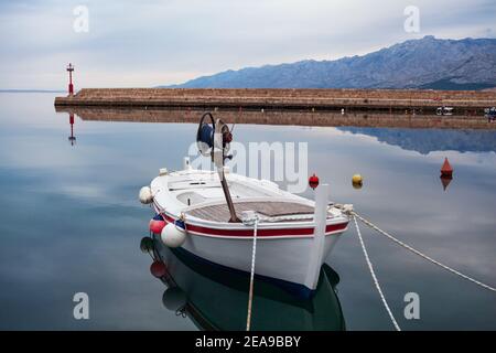 Morgenpanorama mit ruhigem Meer, weißem Fischerboot und Pier mit Leuchtturm und Bergen im Hintergrund. Stockfoto