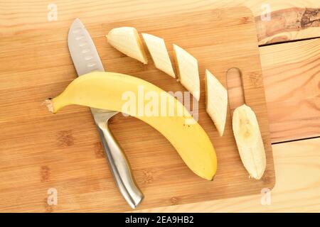 Eine leuchtend gelbe, reife, saftige Bio-Banane und eine geschält und in Scheiben geschnitten, ein Metallmesser - auf einem Schneidebrett aus Bambus, Stockfoto