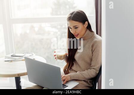 Schöne junge Frau in Freizeitkleidung mit Laptop und genießen Heißgetränk bei der Arbeit im Innenbereich Stockfoto