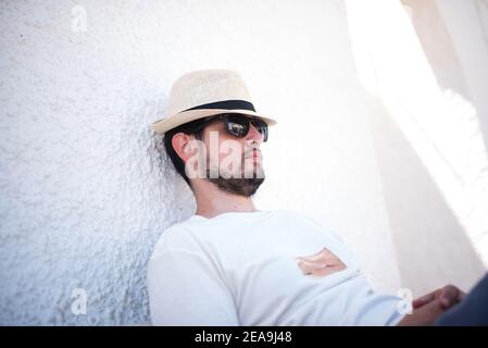 Junger Mann mit Strohhut und Sonnenbrille, der ein Nickerchen im Freien an einer weißen Wand macht Stockfoto
