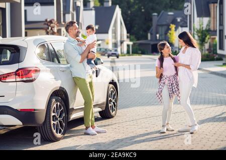 Foto Porträt der Mutter umarmt Tochter, während Vater trägt Sohn Lehnt sich im Sommer auf der Straße an ein weißes Auto Stockfoto