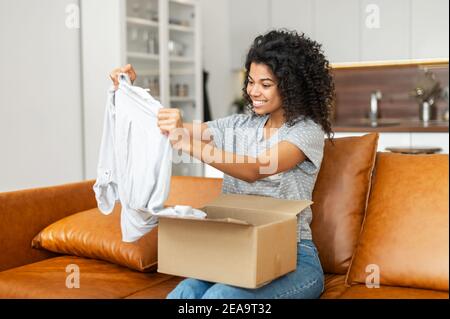 Zufrieden lächelnde junge afroamerikanische Frau auf dem Sofa sitzen Öffnung Paket Karton Box, Auspacken empfangen Geschenk, Artikel aus einem Online-Shop Stockfoto