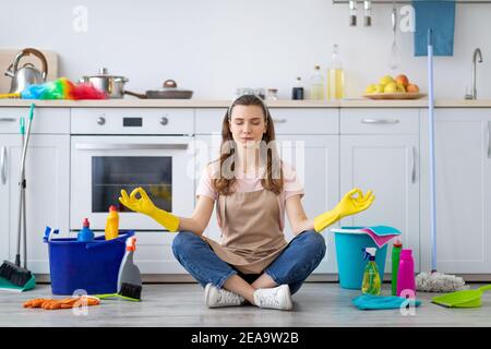 Junge Hausfrau umgeben von Reinigungsmitteln meditieren auf dem Boden in der Küche, Pause von Hausarbeiten Stockfoto