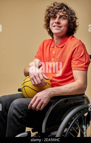 Aufgeschlossener behinderter Kerl hält Basketball und lächelt, er spielt gerne Sportspiele, träumt davon, schneller zu seinen Füßen zu kommen und gibt nicht auf Stockfoto