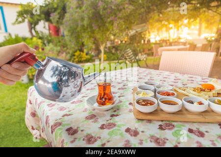 Ein Mann gießt Tee aus einer Teekanne in ein traditionelles türkisches Glas auf der Terrasse eines Restaurants in einem malerischen Garten. Stockfoto