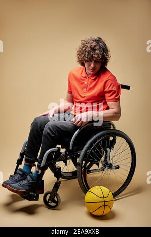 Sportler musste Sport wegen Behinderung aufgeben, er sitzt auf dem Rollstuhl und schaut den Ball mit traurigem Gesichtsausdruck an. Stockfoto