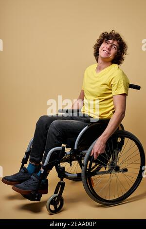 Behinderte fröhlich behinderten Mann berühren Räder und sitzen im Profil, während voran isoliert im Studio, Lächeln. Isoliert auf beigem Hintergrund Stockfoto