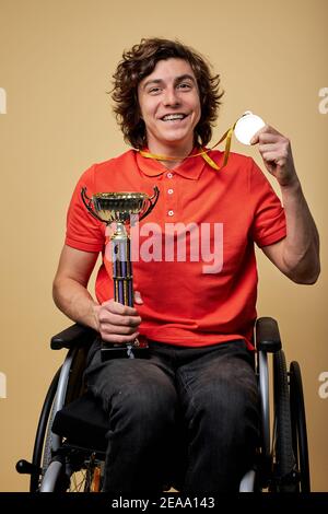 Glücklich behinderten Sportler paralympic im Rollstuhl hält Champion Kelch und Goldmedaillen isoliert auf beigem Hintergrund Stockfoto