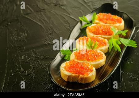 Scheiben frisches Weißbrot mit Butter und rotem Kaviar  mit Petersilie auf der schwarzen Schale und auf dem schwarzen Hintergrund. Stockfoto