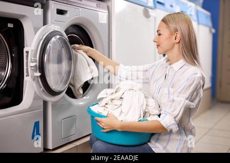 Frau, die Wäsche tut - weiße Kleider aus der Waschmaschine herausstellen, sie in Korb, Waschbecken setzen. In Waschhaus Stockfoto