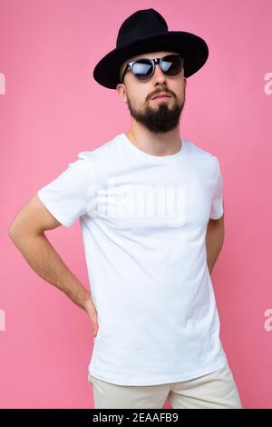 Vertikale Foto-Aufnahme von schönen Brünette erwachsenen Mann mit Bart Tragen lässige weiße T-Shirt für Mockup schwarzen Hut und stilvoll Sonnenbrillen isoliert über Stockfoto