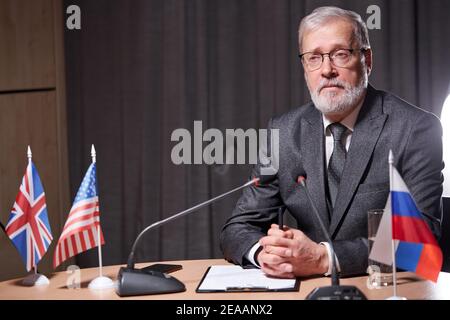 Senior Geschäftsmann sitzt mit Mikrofon in modernen Sitzungssaal während der Sitzung mit Geschäftspartnern, gutaussehende Mann in Brillen selbstbewusst sitzen, Stockfoto