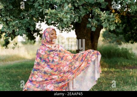 Muslimische schwarze Frau afrikanische Ethnizität getragen traditionellen bunten Hijab lächelt und schaut rechts unter grünen Baum. Geschäft für schwarze Frau. Stockfoto