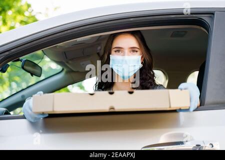 Schöne kaukasische Mädchen mit grünen Augen schaut in die Kamera und lächelt. Junges Mädchen hält Pizza im Auto und arbeitet als Kurier in Lieferfirma bei quaranti Stockfoto