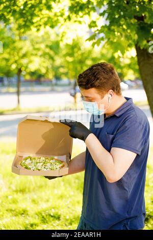 Kurier in medizinische Maske und Handschuhe hält Pizza in den Händen und sieht Pizza mit grünem Salat, Tomaten, Käse. Vertikales Foto für Werbung in sozialen Medien Stockfoto