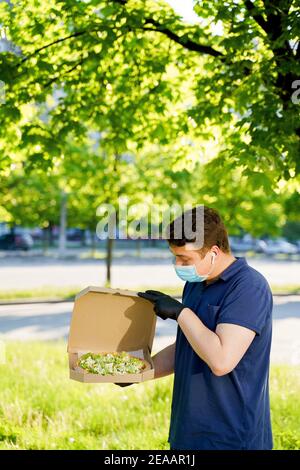 Kurier in medizinische Maske und Handschuhe hält Pizza in den Händen und sieht Pizza mit grünem Salat, Tomaten, Käse. Vertikales Foto für Werbung in sozialen Medien Stockfoto