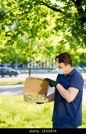 Kurier in medizinische Maske und Handschuhe hält Pizza in den Händen und sieht Pizza mit grünem Salat, Tomaten, Käse. Vertikales Foto für Werbung in sozialen Medien Stockfoto