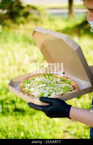 Kurier in medizinische Maske und Handschuhe hält Pizza in den Händen und sieht Pizza mit grünem Salat, Tomaten, Käse. Vertikales Foto für Werbung in sozialen Medien Stockfoto