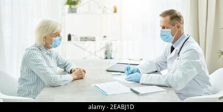 Arzt in medizinischer Maske sitzt am Tisch und spricht Mit einer alten Dame Stockfoto